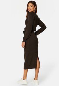 Abito a maglia, con coste, a maniche lunghe in marrone scuro con spacco laterale. Abbinato a sneakers bianche. Scollo rotondo semplice, design aderente.