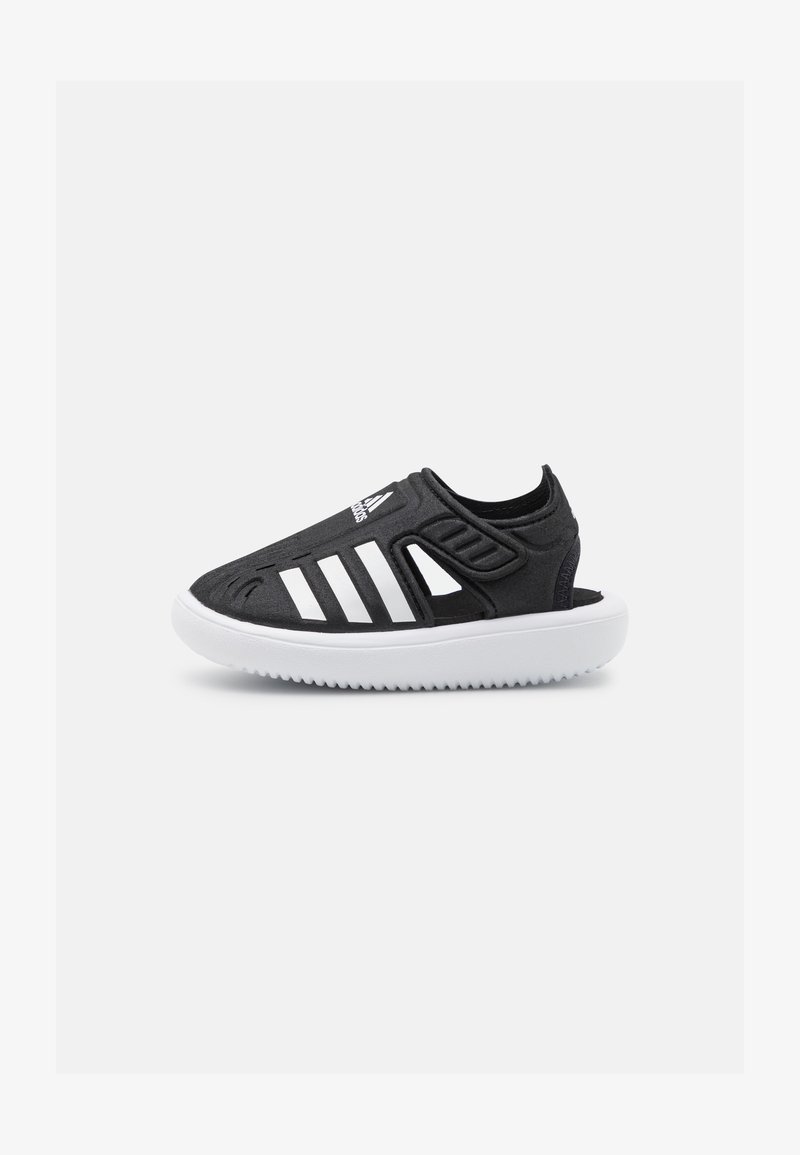 Black athletic shoe with white stripes, featuring a textured upper and open design. Thick white sole with a ribbed pattern.