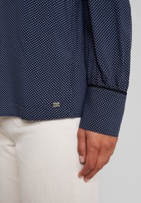 Tummansininen pilkullinen pusero, rento malli, pitkät hihat ja mustat kalvosimet. Puserossa on pieni metallinen logo lähellä helmaa.