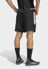 Άτομο που φοράει μαύρες αθλητικές βερμούδες με άσπρες ρίγες, μαύρες κάλτσες και μαύρα ποδοσφαιρικά παπούτσια με φωσφορίζοντα κίτρινα στοιχεία, στέκεται με τα χέρια στη μέση.