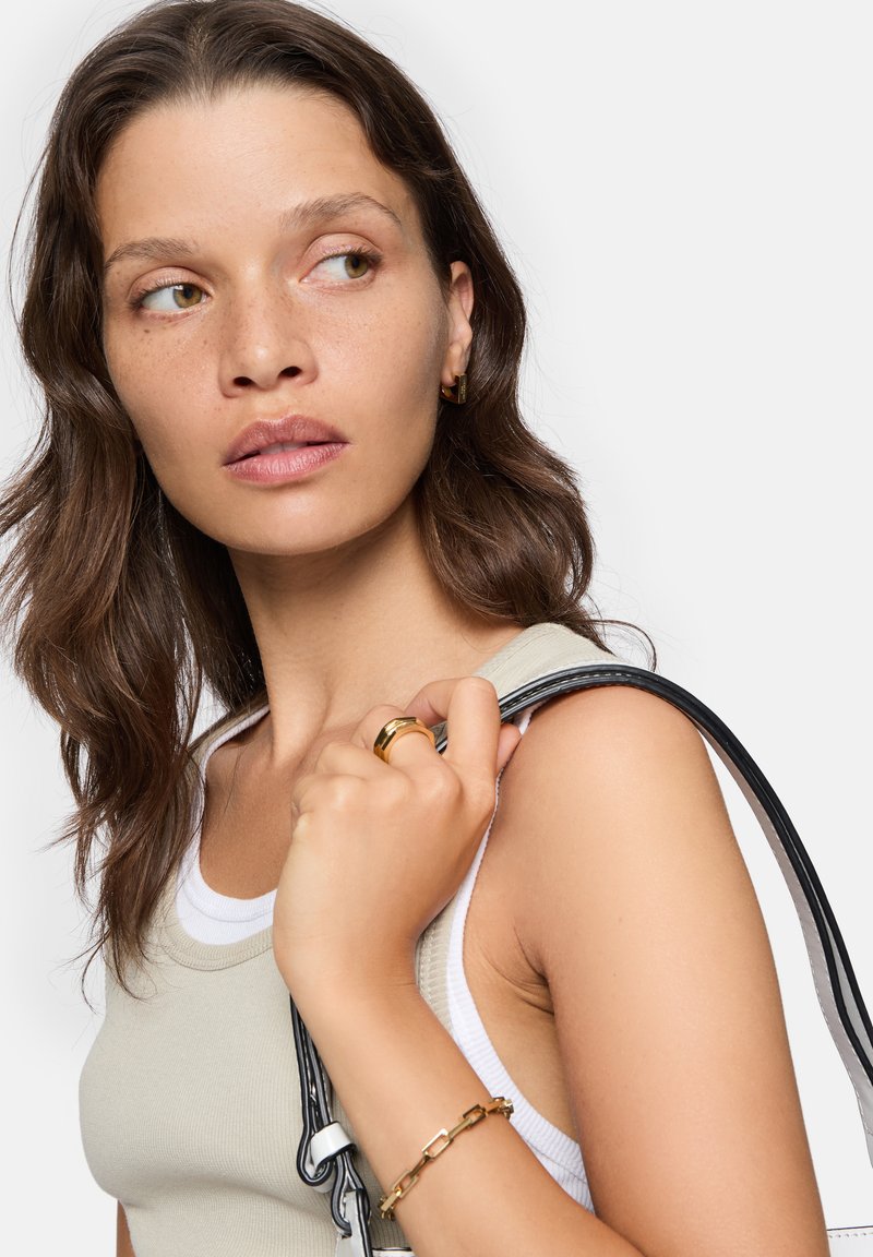 Beige tanktop over een wit shirt, met een zwarte strap, en een gouden ring en armband. Haar is golvend en bruin. Gelaat met sproeten.