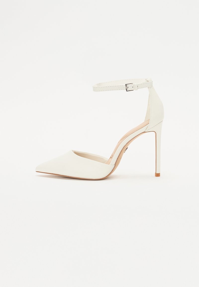 Chaussure blanche à talon haut avec bout pointu et bride à la cheville avec boucle argentée, présentée sur un fond blanc uni.