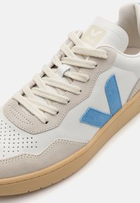 Hvit og grå sneaker med en blå fløyels "V"-logo, teksturert skinn og fløyel overdel, beige snørebånd og gummisåle. Perforert tåhette.