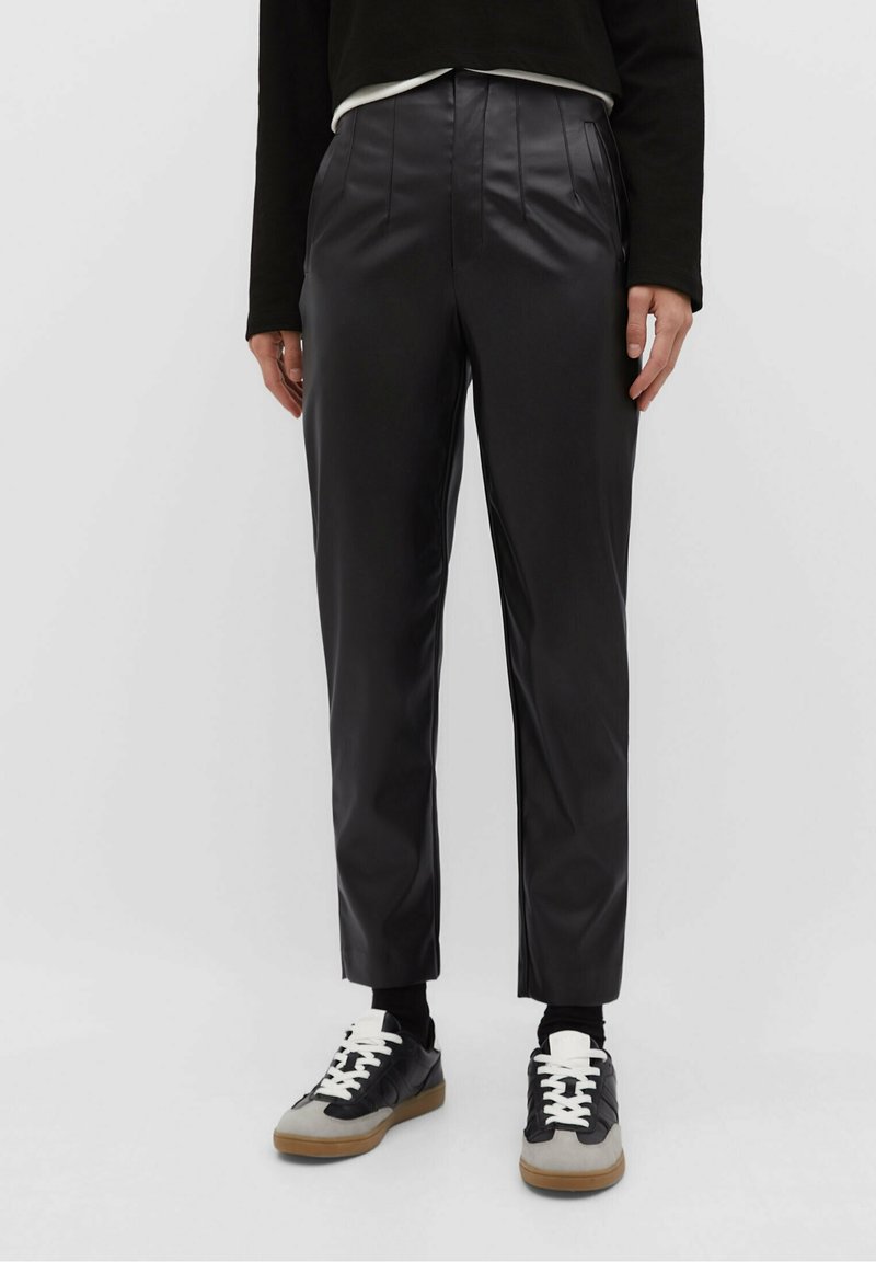 Zwarte neplederen broek met voorplooien en een taps toelopend been, gecombineerd met zwart-grijze sneakers met witte veters en rubberen zolen.
