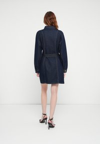 Robe chemise en denim bleu foncé, ceinturée à la taille, avec de longues manches bouffantes et des poignets boutonnés. Portée avec des talons noirs et des sangles à pois.