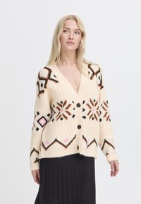 Cárdigan crema con patrones geométricos en marrón, rosa y negro; presenta un escote en V, cinco botones y un punto texturizado.