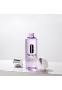 Démaquillant Clinique dans une bouteille transparente violette avec du texte noir, accompagné d'une boule de coton et d'un capuchon argenté sur une surface blanche.