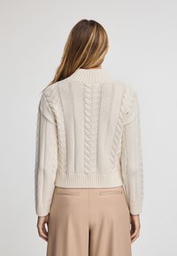 Krämfärgad stickad tröja med ribbad krage och texturerade flätmönster på ryggen. Kort design med långa ärmar.