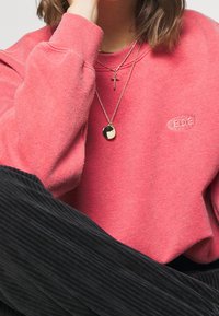 Koralfleece sweatshirt med liten logotyp, lager av guldhalsband med ett kors och en texturerad berlock. Svarta ribbade byxor.