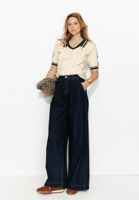 Polo beige à manches courtes avec un col foncé, associé à un pantalon en denim large et foncé et des chaussures marron à enfiler. Tient une pochette à imprimé léopard.