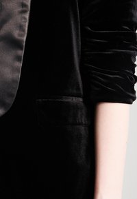 Černé sametové sako se saténovým klopem, s jednou náprsní kapsou a nařasenými rukávy. Měkká textura, zřejmé elegantní designové detaily.
