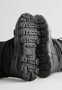 Zwarte schoenen met robuuste rubberen zolen met diepe noppen en ingewikkelde patronen, ontworpen voor grip en duurzaamheid.