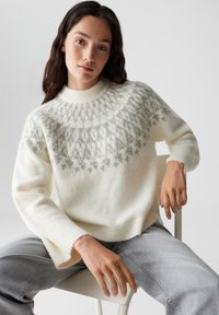 Maglione bianco in lana con un design a motivo grigio sullo scollo, colletto e maniche a coste. Abbinato a jeans grigi chiari.