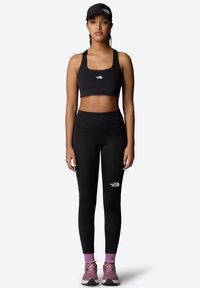 Sujetador deportivo negro y leggings de tiro alto, ambos con el logo de North Face. La modelo lleva una gorra negra y zapatillas con acentos en morado.