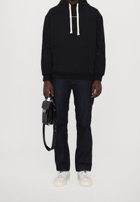 Persona vestida con una sudadera negra con cordones blancos, jeans oscuros, zapatillas blancas y sosteniendo un pequeño bolso negro en una mano.