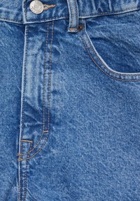 Gros plan de la partie avant haute d’un jean en denim bleu montrant un bouton en métal, une boucle de ceinture, une poche et des coutures orange.