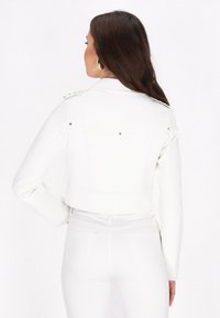 Giacca in ecopelle bianca corta con maniche lunghe, dettagli con bottoni sulle spalle e tasche posteriori, abbinata a jeans bianchi aderenti.