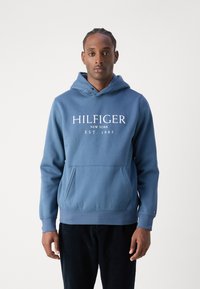 Uomo che indossa un hoodie blu Hilfiger con tasca frontale e il testo "HILFIGER NEW YORK EST. 1985" in piedi contro uno sfondo semplice.