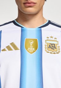 Man som bär Argentinas fotbollströja med Adidas-logotyp, FIFA världsmästarna 2022-märke och Argentinska fotbollsförbundets emblem.