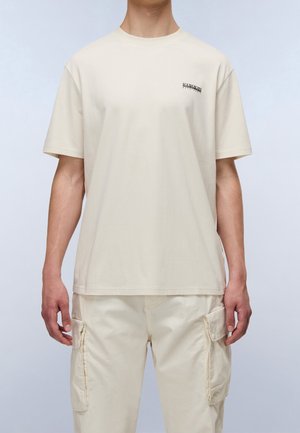 Person iført en ensfarvet lysebeige kortærmet t-shirt med lille logo på brystet og matchende cargobukser med sidelommer, stående oprejst.