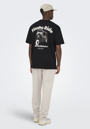 Uomo con t-shirt nera con grafica di tuk-tuk e testo "Fiesta Ride", pantaloni beige, cappellino beige e scarpe da ginnastica di colore chiaro, in piedi di schiena.