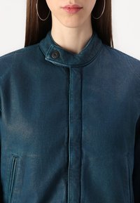 Chaqueta de mezclilla azul con cuello alto, cierre frontal de botones a presión y bolsillos laterales; presenta un acabado texturizado y ligeramente brillante.