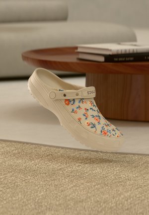 Sabot beige à motif floral et bride de talon réglable flottant au-dessus d'un tapis beige texturé, table en bois et livres en arrière-plan flou.