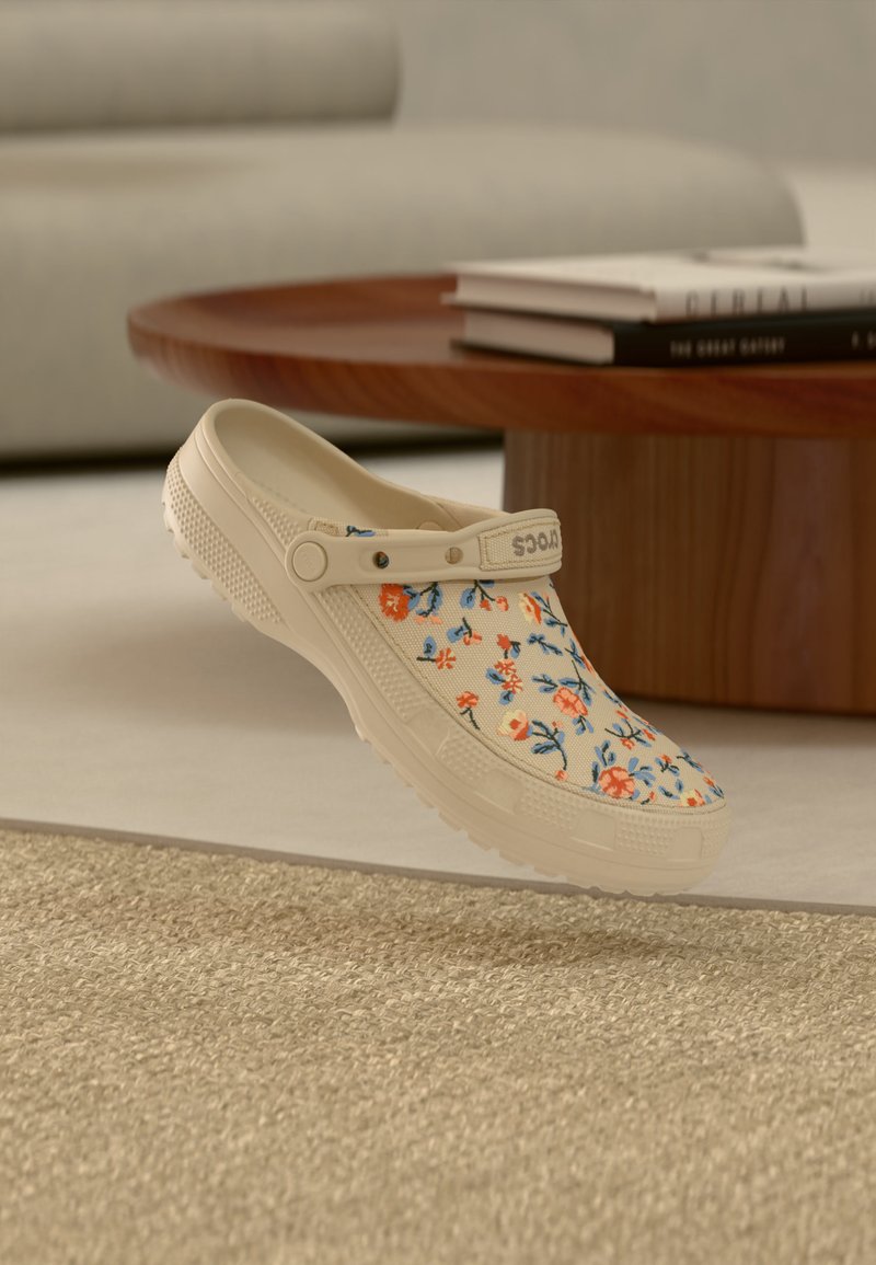 Sabot beige à motif floral et bride de talon réglable flottant au-dessus d'un tapis beige texturé, table en bois et livres en arrière-plan flou.