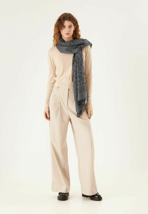 Grijze sjaal met patroon gewikkeld om een beige trui, gedragen met beige wijde broek en zwarte schoenen, staande tegen een witte achtergrond.