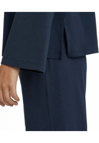 Marineblaues Baumwolloberteil mit weiten Ärmeln und seitlichen Schlitzen. Sichtbare Handdetails, die eine weiche Textur und einen entspannten Schnitt zeigen.