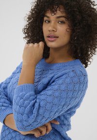 Blå strikket sweater med et struktureret mønster, brede ribbede manchetter og rund halsudskæring. Stofmaterialet er let og luftigt.
