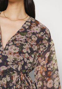 Mekko, jossa on kukkakuosi, valmistettu läpikuultavasta, ryppyisestä kankaasta. Siinä on V-mallinen kaula-aukko, pitkät hihat ja solmittava vyötärö, tummalla taustalla ja vaaleanvärisillä kukilla.