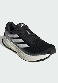 Chaussure de course noire avec une tige en mesh texturé, logo à trois bandes blanches et une semelle rembourrée. Elle dispose de lacets et d'un col rembourré.
