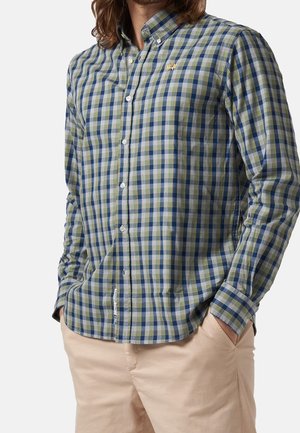 Mann trägt ein grün-blau-weiß kariertes Hemd mit Knöpfen und beige Hose, Hände in den Taschen, vor einfachem hellem Hintergrund.