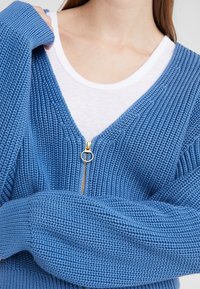 Blå stickad cardigan med V-hals, med guld dragkedja och ribbade ärmslut. Bärs över en vit T-shirt. Texturerat tyg.