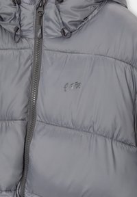 Veste matelassée grise avec capuche, fermeture éclair devant, cordons de serrage et logo manuscrit discret sur la poitrine.