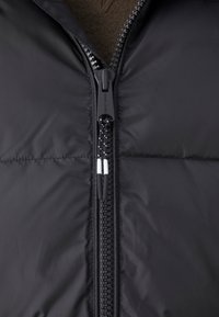 Sort pufferjakke med lynlås foran og mærket på pull tab, tyk polstring og glat ydre overflade. Ingen synlige mønstre eller detaljer.