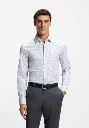 Camicia bianca con piccoli motivi blu e neri, colletto con bottoni, maniche lunghe, texture minima, abbinata a pantaloni grigi e cintura nera.