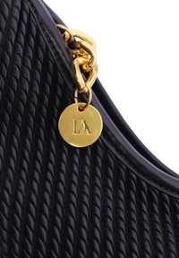 Bolso negro con superficie texturizada, que presenta una cadena dorada y una etiqueta redonda con un logotipo en relieve. Bordes lisos y curvados.