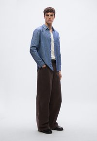 Svetlo modra srajca z gumbi in žepom, združena s temno rjavimi širokimi hlačami in črnimi čevlji, z ohlapnim krojem in gladko teksturo.