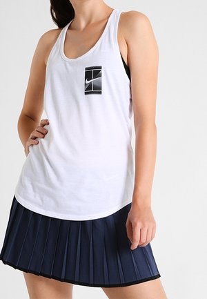 Femme portant un haut sans manches blanc Nike avec logo noir et jupe plissée bleu marine, posant avec une main sur la hanche.