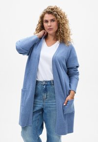 Cardigan lungo azzurro chiaro in morbido tessuto, con maniche a coste e due tasche frontali, indossato sopra una maglietta bianca con scollatura a V.