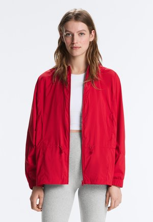 Žena oblečená v červenej bunde na zips cez biely crop top a sivé legíny, stojaca pred bielym jednoduchým pozadím.