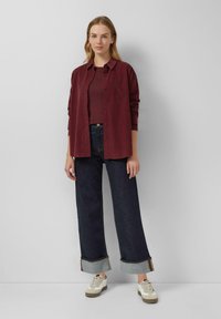 Bordeaux knoop-overhemd over een kastanjebruine top, gecombineerd met wijde donkerdenim jeans met opgerolde boorden en retro-stijl witte sneakers.