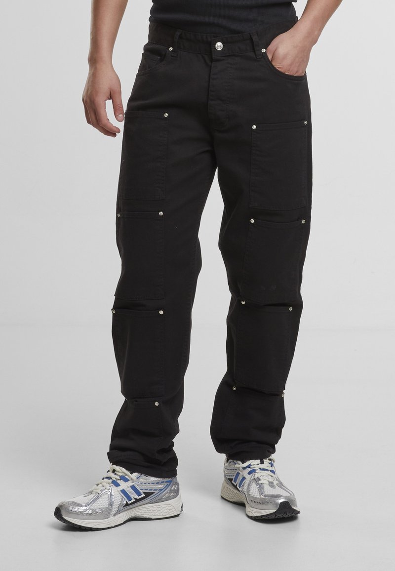 Personne portant un pantalon cargo noir avec de multiples poches et des boutons argentés, associé à des baskets de sport blanches et bleues.