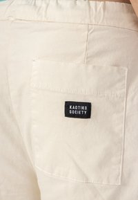 Biele nohavice s vreckom na zadnej strane, na ktorom je čierna nálepka s bielym nápisom "KAOTIKO SOCIETY". Bavlnený materiál s hladkou textúrou.