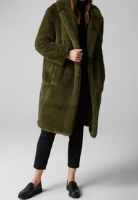 Oliven grøn kunstpelsfrakke med bred krave, dobbeltradet design, store knapper og sidelommer. Båret over sort tøj og loafers.