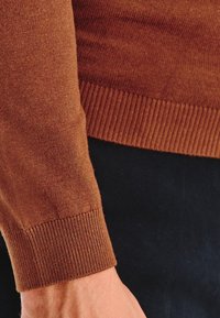 Brązowy sweter z długim rękawem, mankiet z detalami w pr�ążki na ramieniu osoby na ciemnym tle.