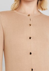 Beżowy dzianinowy kardigan z okrągłym dekoltem i złotymi guzikami z przodu. Gładka faktura i dopasowany fason.