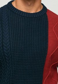 Maglione a girocollo lavorato a maglia con sezioni blu scuro e rosse, con texture a treccia sul lato blu e tessuto liscio sul lato rosso.